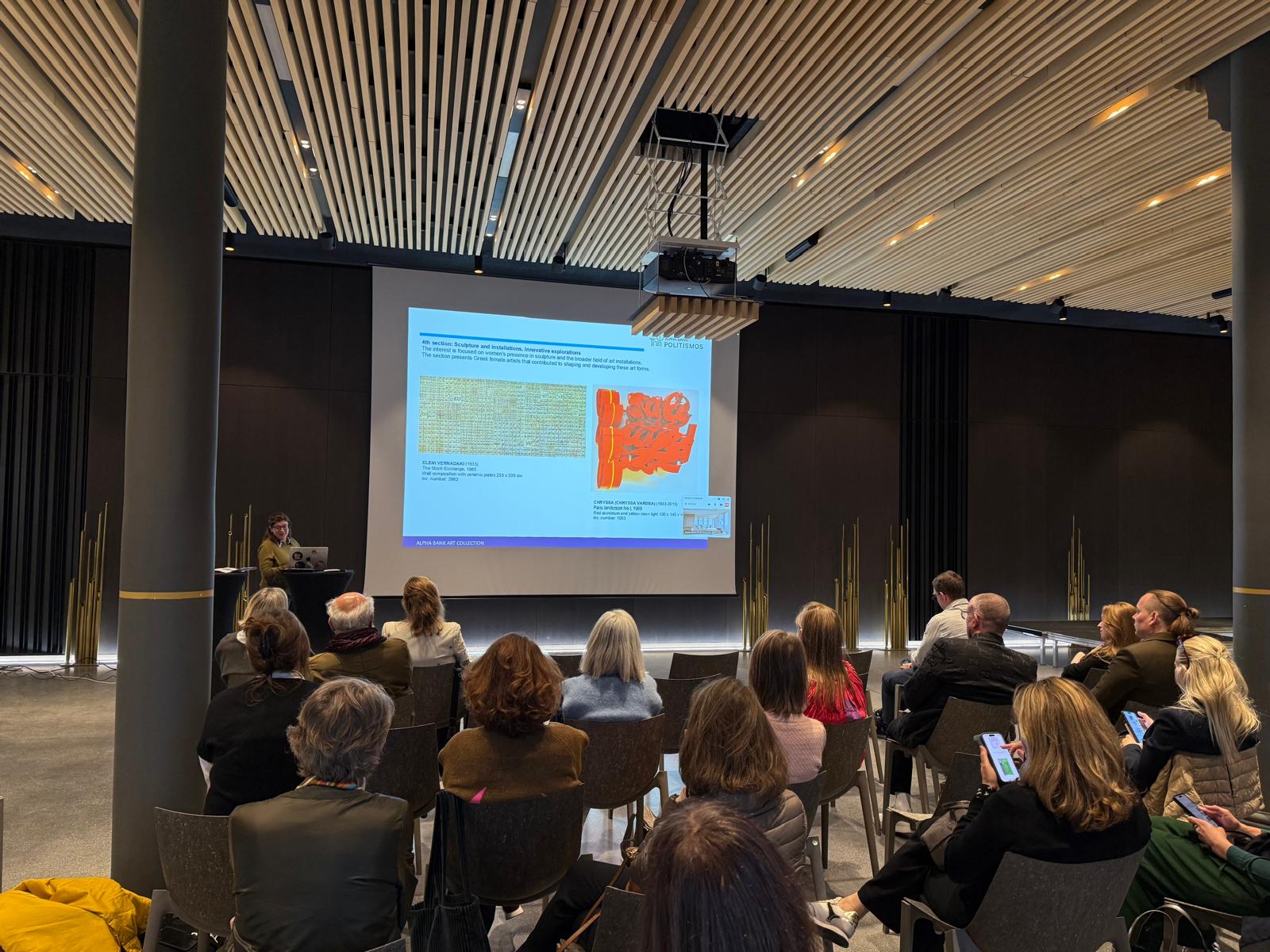 Ομιλήτρια μπροστά από laptop και δίπλα από οθόνη προβολής παρουσιάζει σε κοινό με άντρες και γυναίκες. Στιγμιότυπο από την παρουσίαση