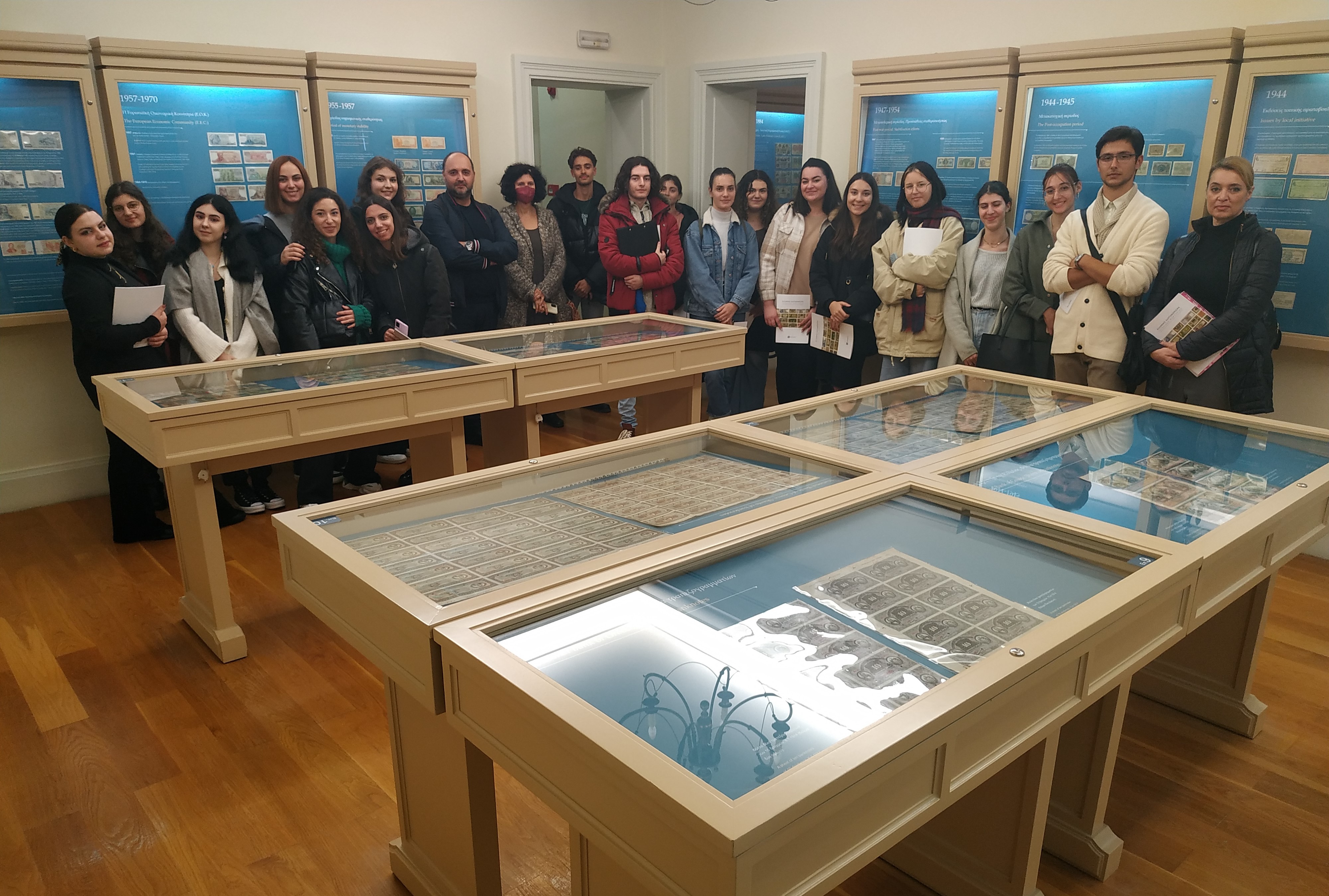 Students and academic staff standing behind display cases containing coins in a museum hall.  The Banknote Museum of Ionian Bank on Corfu partners with the Ionian University