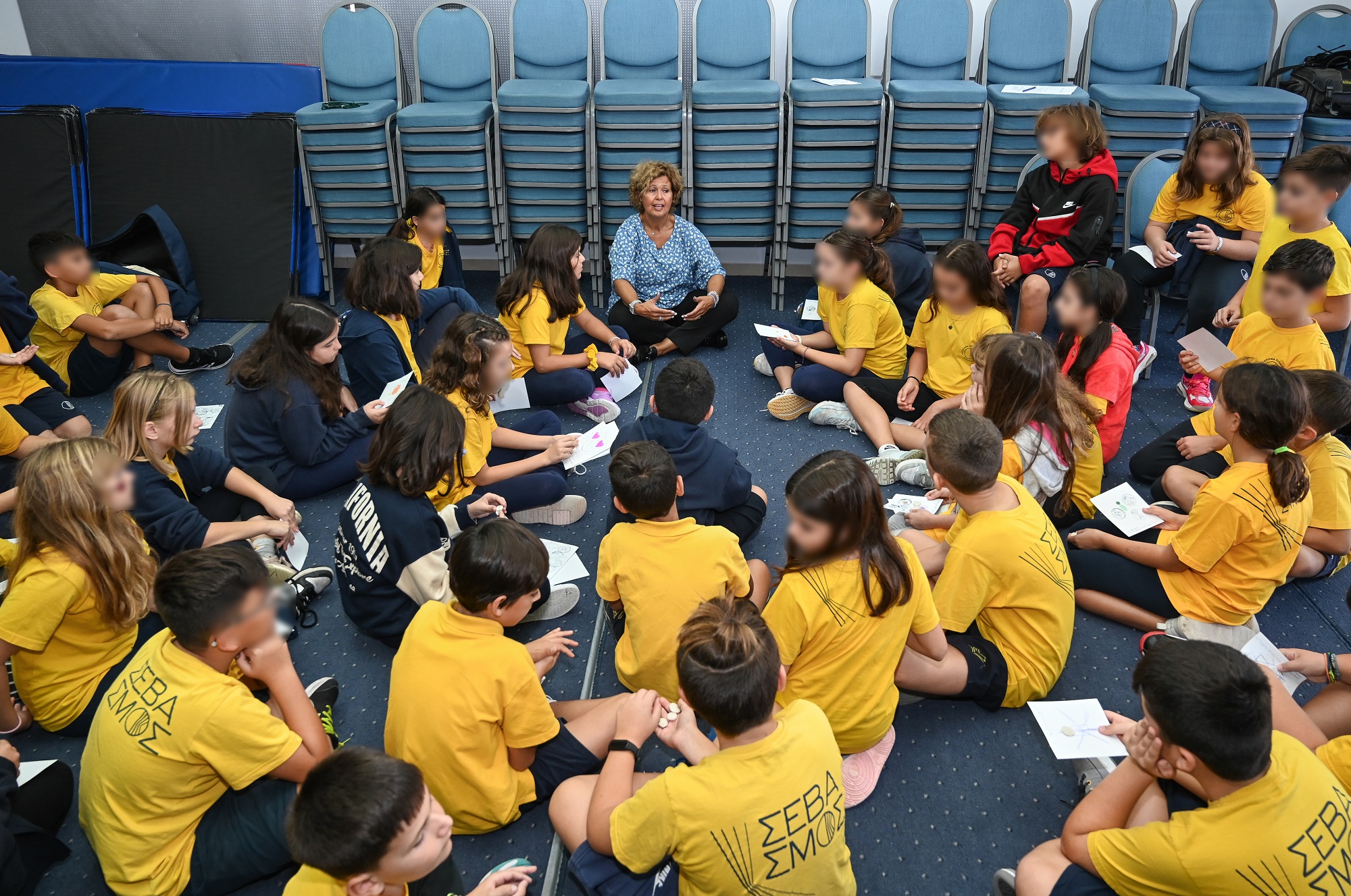 Η κυρία Τσαγκάρη καθισμένη στο πάτωμα εξηγεί σε μαθητές και μαθήτριες που κάθονται γύρω της.  Στιγμιότυπο από την παρουσίαση