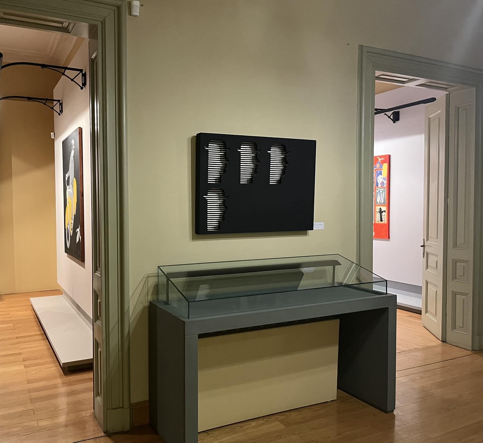Glass display case in front of olive-green wall. Above the work Untitled, acrylic on plywood assemblage.  The exhibition venue at the Municipal Art Gallery of Thessaloniki