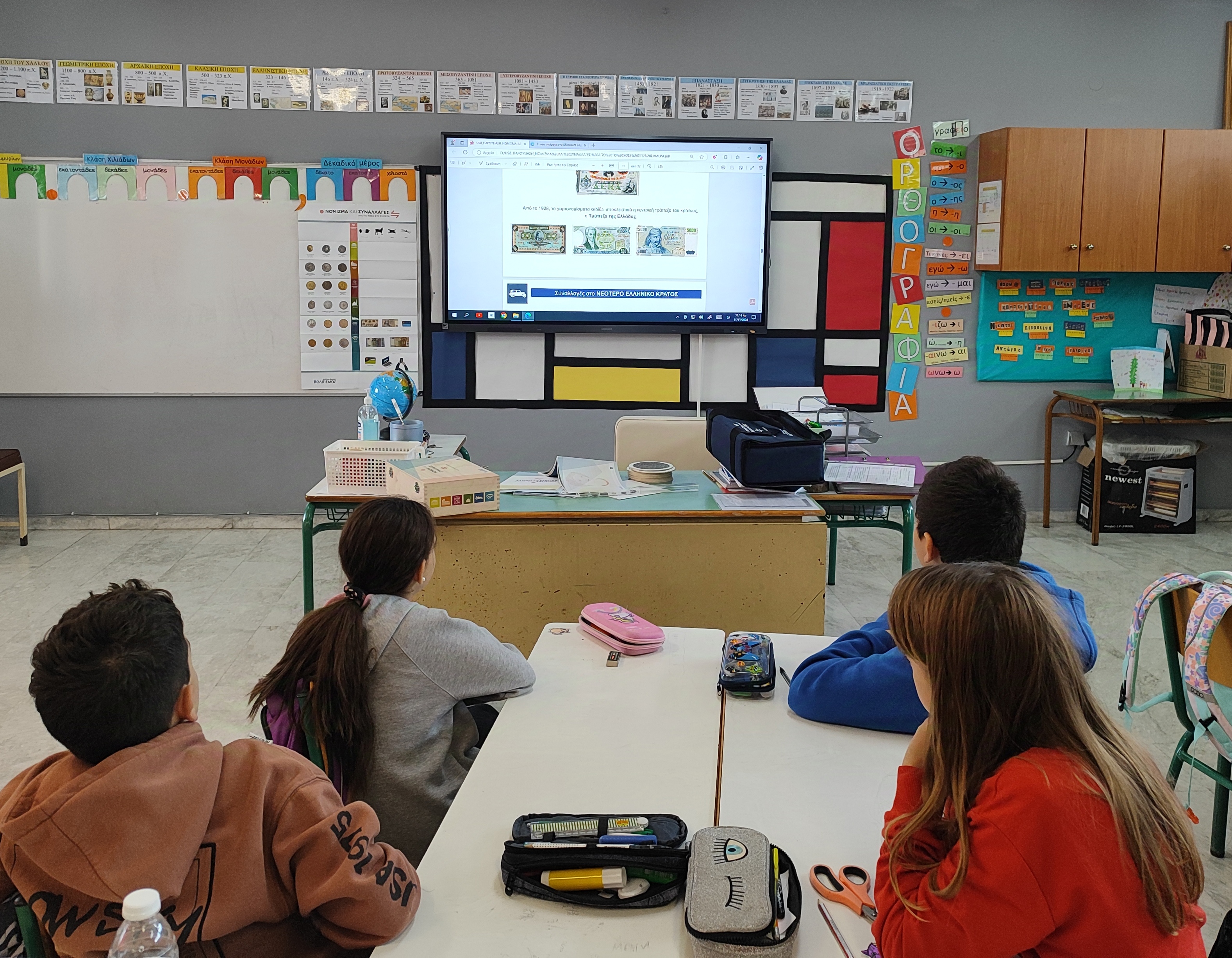 2 boys and 2 girls watching a presentation of the “Currency and Transactions” programme in a classroom. Presentation on an interactive board