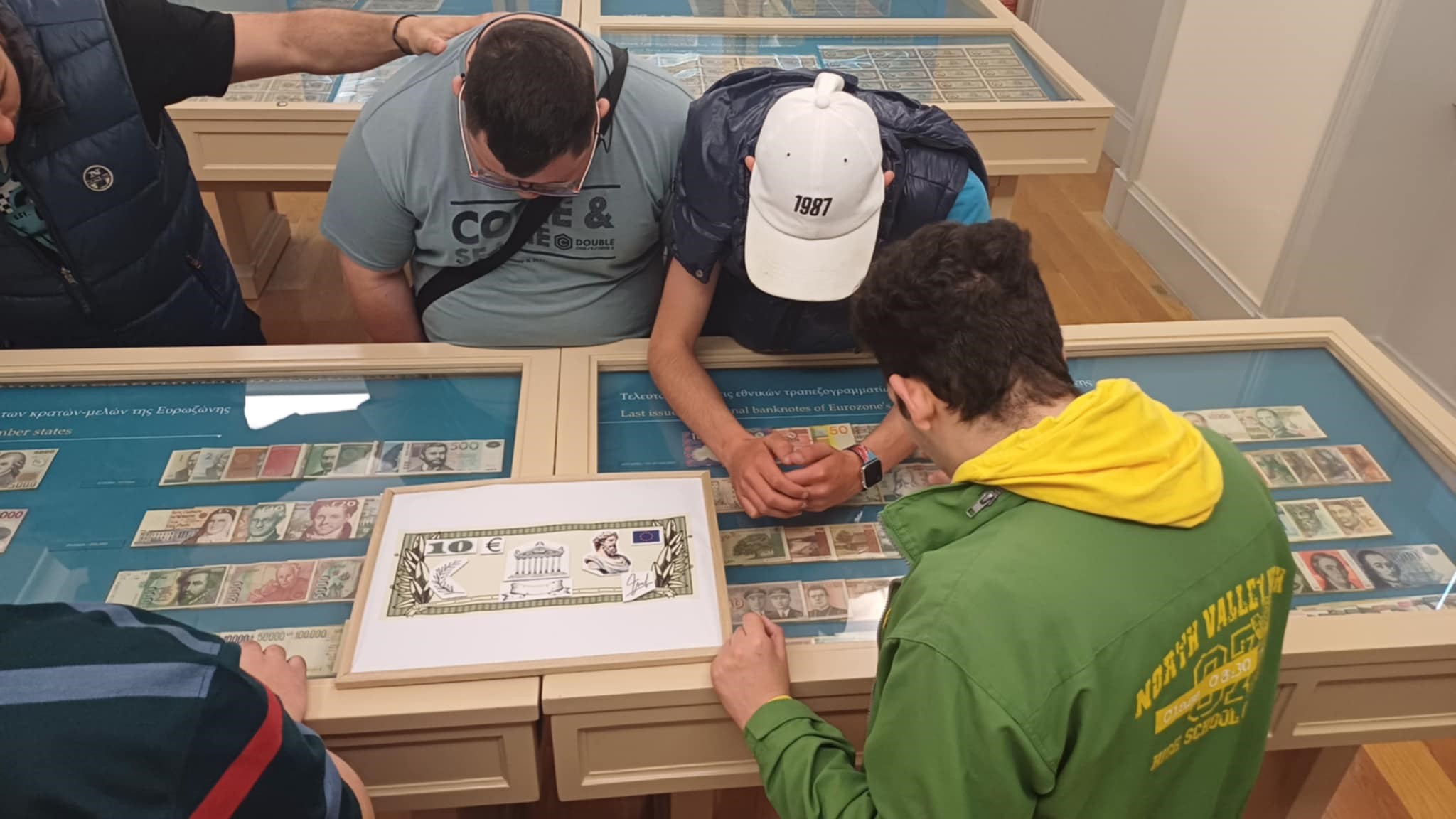 Visitors from the “Dimotiki Folia” KDAP MEA gathered around a display case with banknotes. On it, an enlarged replica. Highlight from educational activity