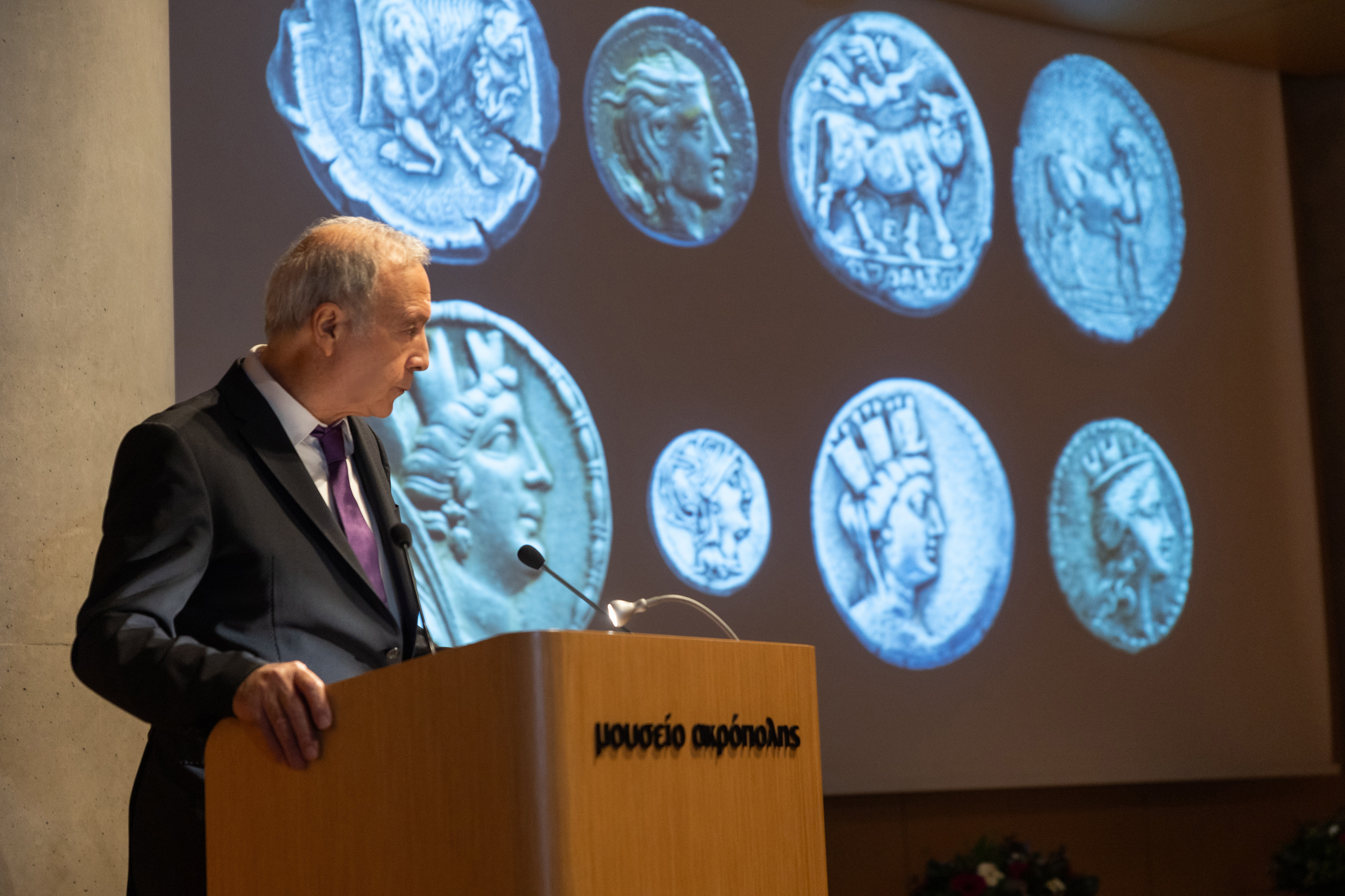 The Acropolis Museum General Manager Nikolaos Stampolidis standing in the podium. A projection of coins in the back. Highlight from the exhibition opening