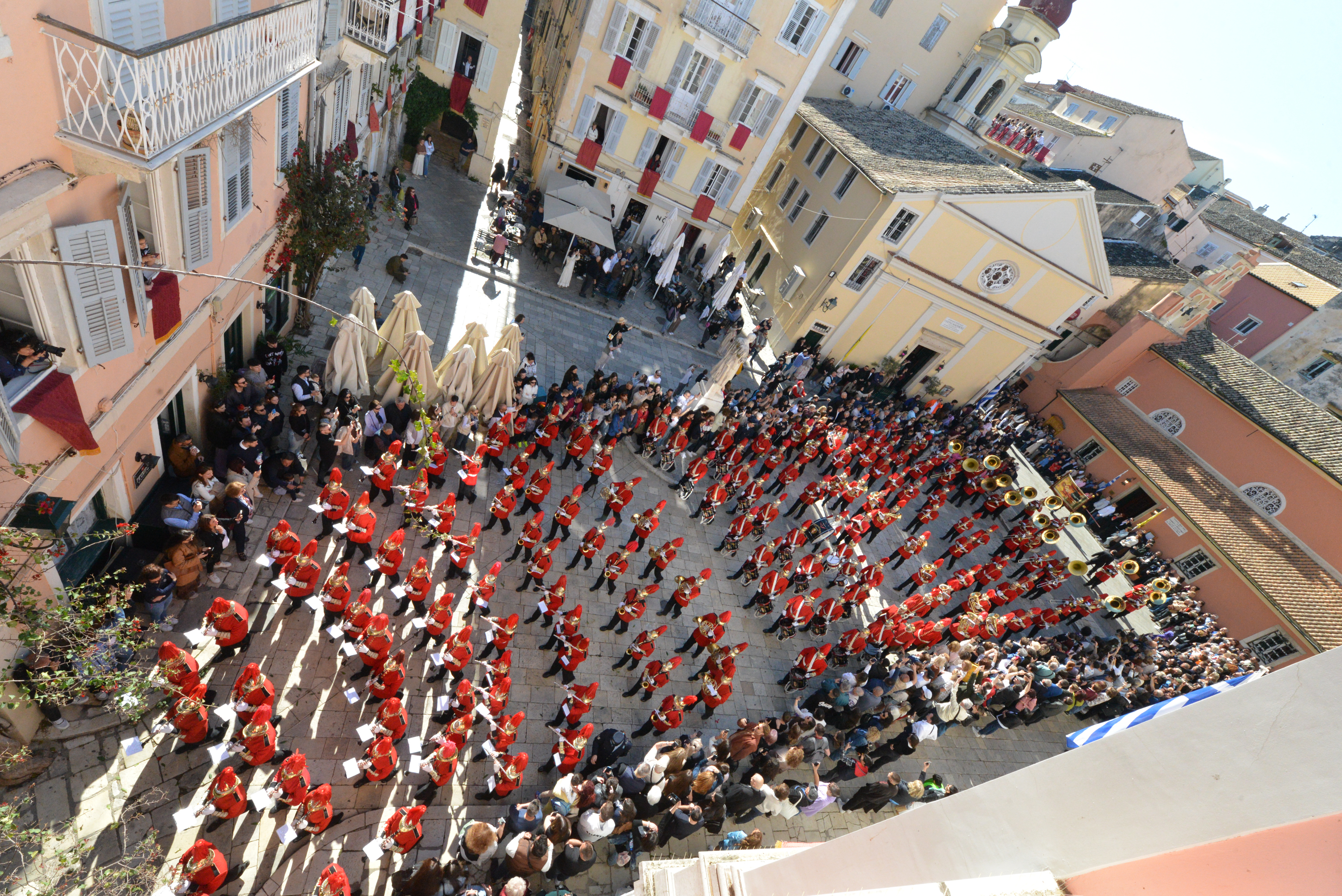 Musicians of the Kapodistrias Philharmonic Association in red uniforms and hats marching through the crowded square. The Kapodistrias Philharmonic Association